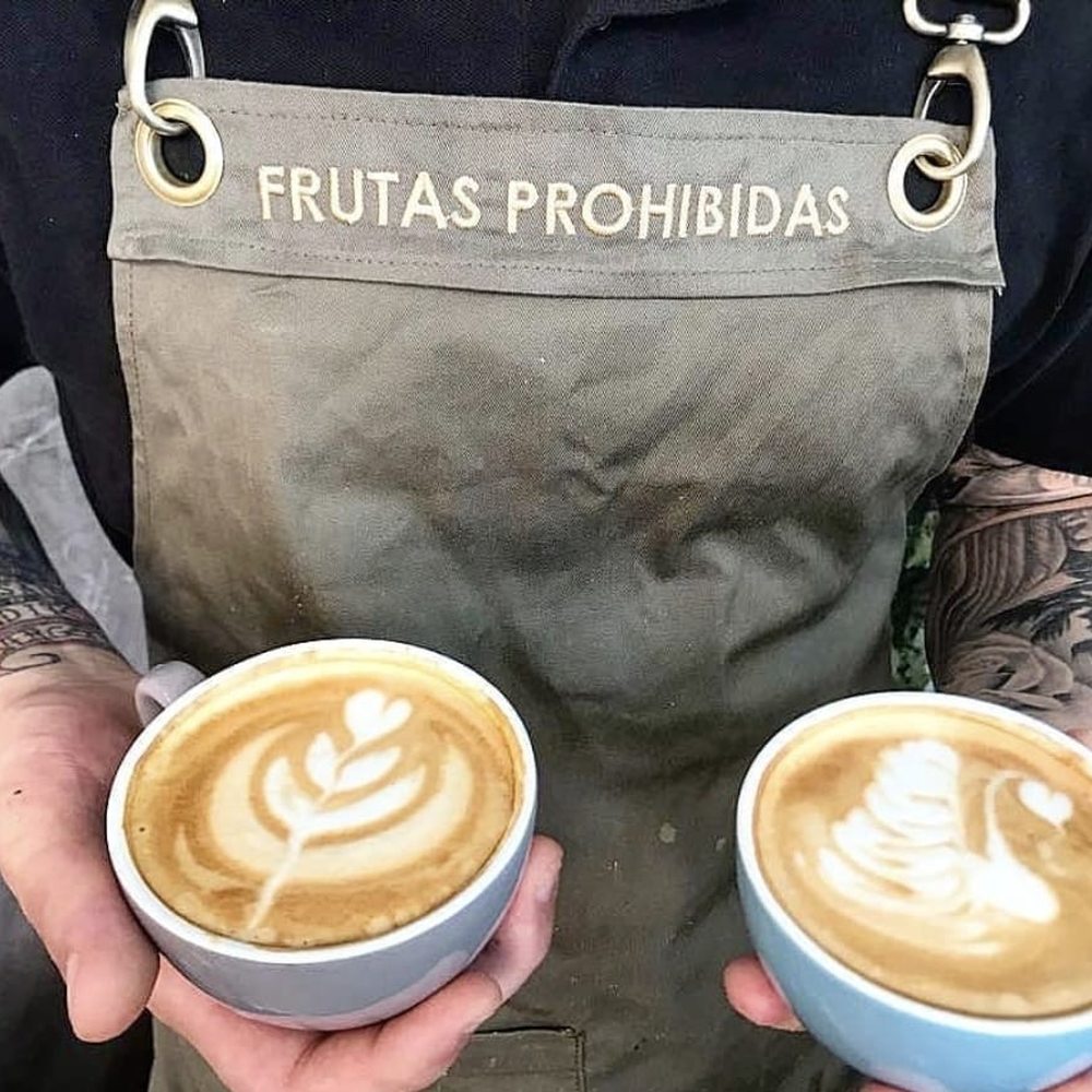 Barista de Frutas Prohibidas con dos cafés con latte art y el delantal de la casa