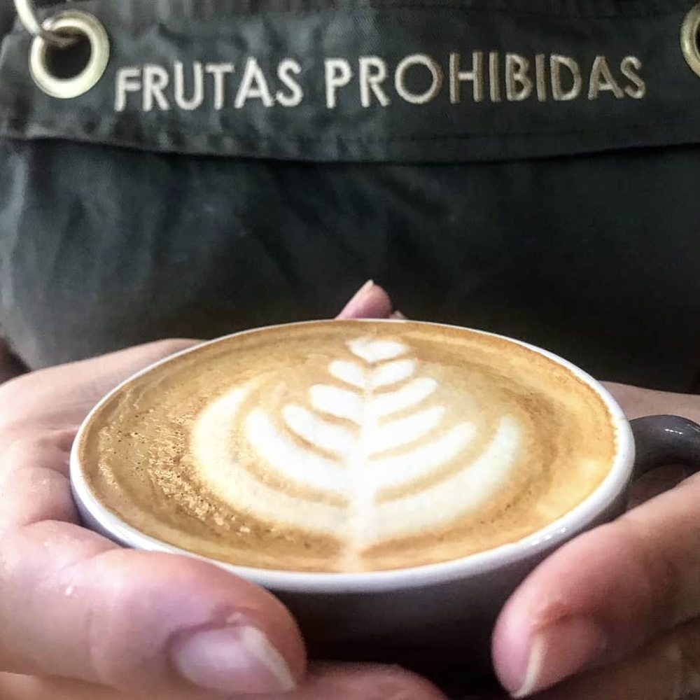 Barista de Frutas Prohibidas con dos cafés con latte art y el delantal de la casa